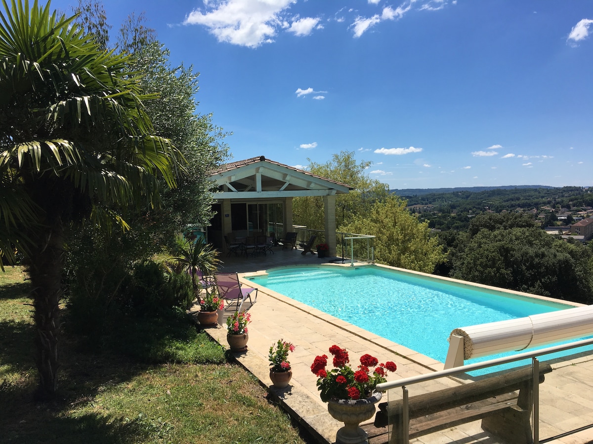 The image features a serene outdoor scene showcasing a spacious swimming pool surrounded by lush greenery. A covered patio area with seating is visible, providing a view of the landscape. Potted flowers add vibrant color to the setting under a clear blue sky.