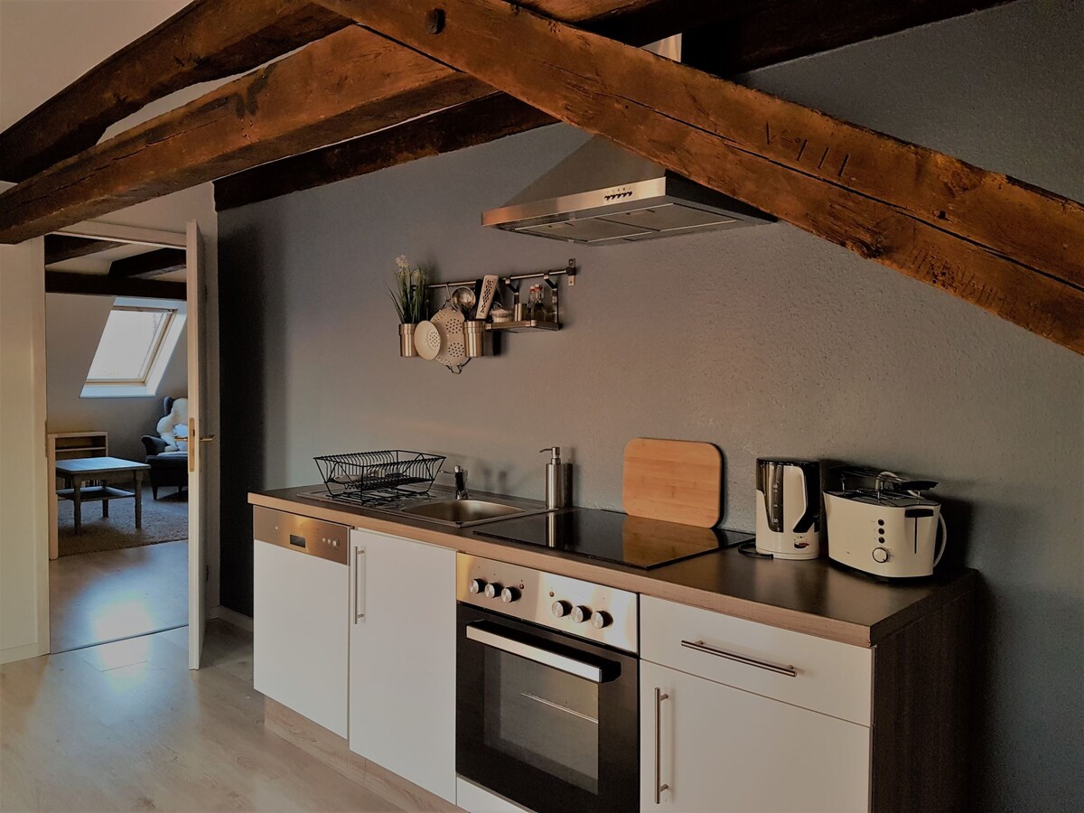 A modern kitchen area features sleek cabinetry, a cooktop, and an oven beneath wooden beams. A kettle and a toaster are positioned on the countertop, complemented by a wall shelf holding decorative items. The adjoining living space is visible through an open door.