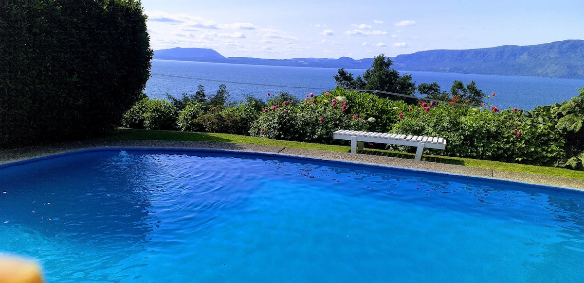 A serene swimming pool glistens under the sunlight, reflecting the clear blue sky. The surrounding lush greenery and blooming flowers frame the area, enhancing the view of the tranquil lake and distant mountains in the background.