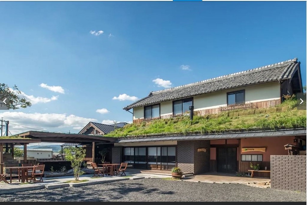 The exterior of the renovated traditional Japanese house is visible, featuring a thatched roof and wooden elements. A spacious patio area with outdoor seating is present, surrounded by greenery and gravel. The setting is complemented by a clear sky and light clouds.