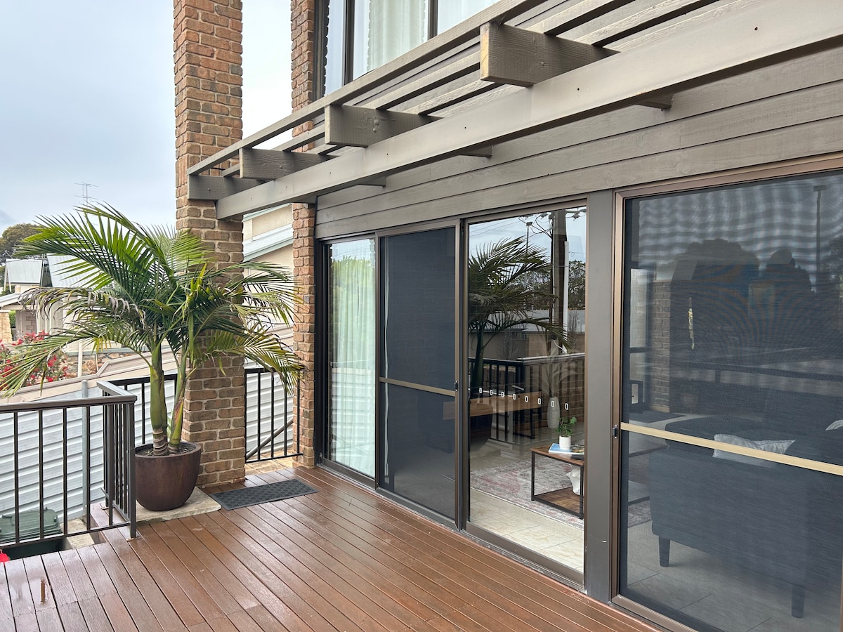 A spacious outdoor balcony features a wooden deck and is bordered by a tall potted palm. Large glass sliding doors lead into the living area, while the surrounding neighborhood is visible in the background.