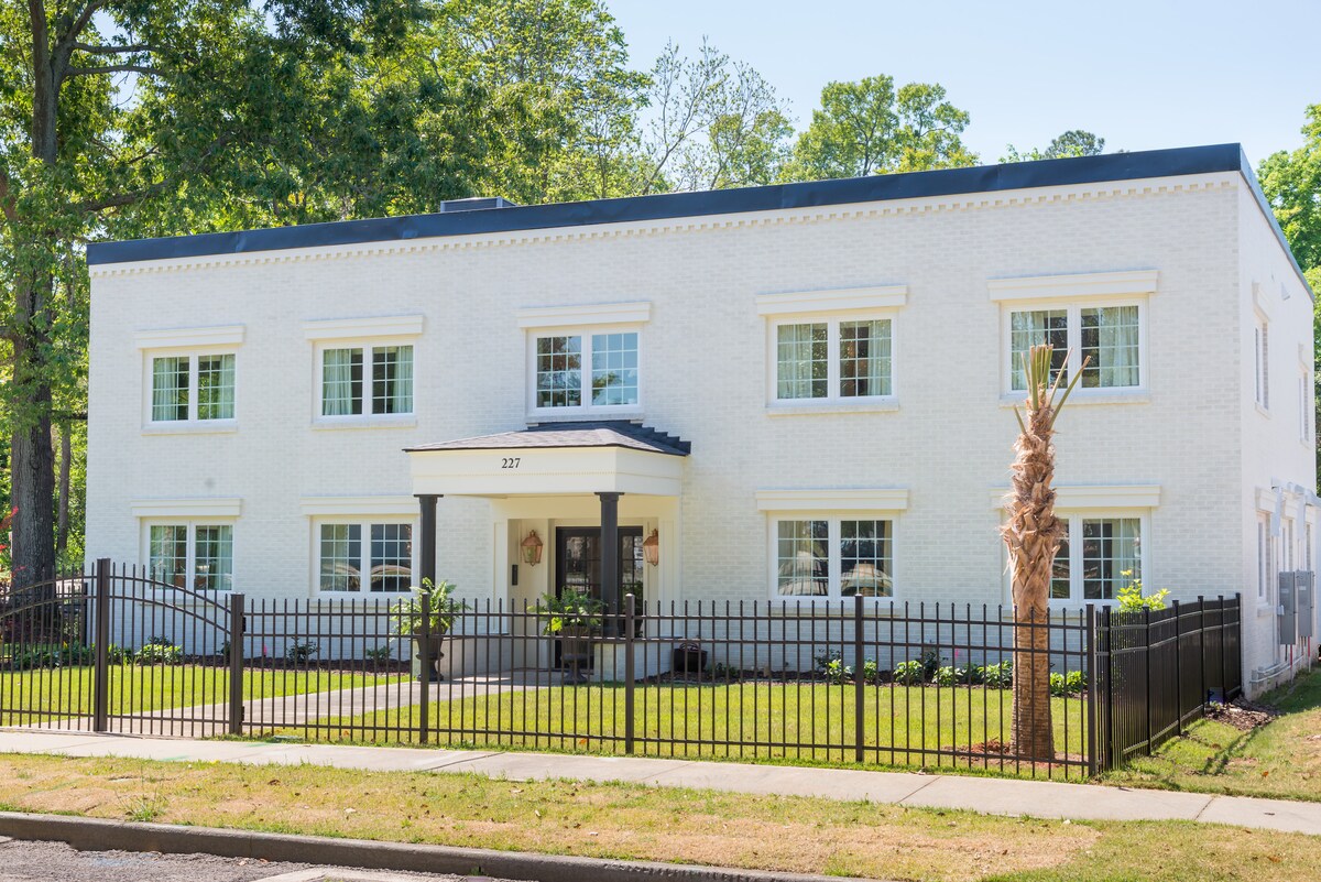The exterior of a renovated building is presented, featuring a white facade and a central entrance under a small roof. Large windows allow light to fill the interior, while palm trees and landscaping enhance the property. A black wrought iron fence outlines the area.
