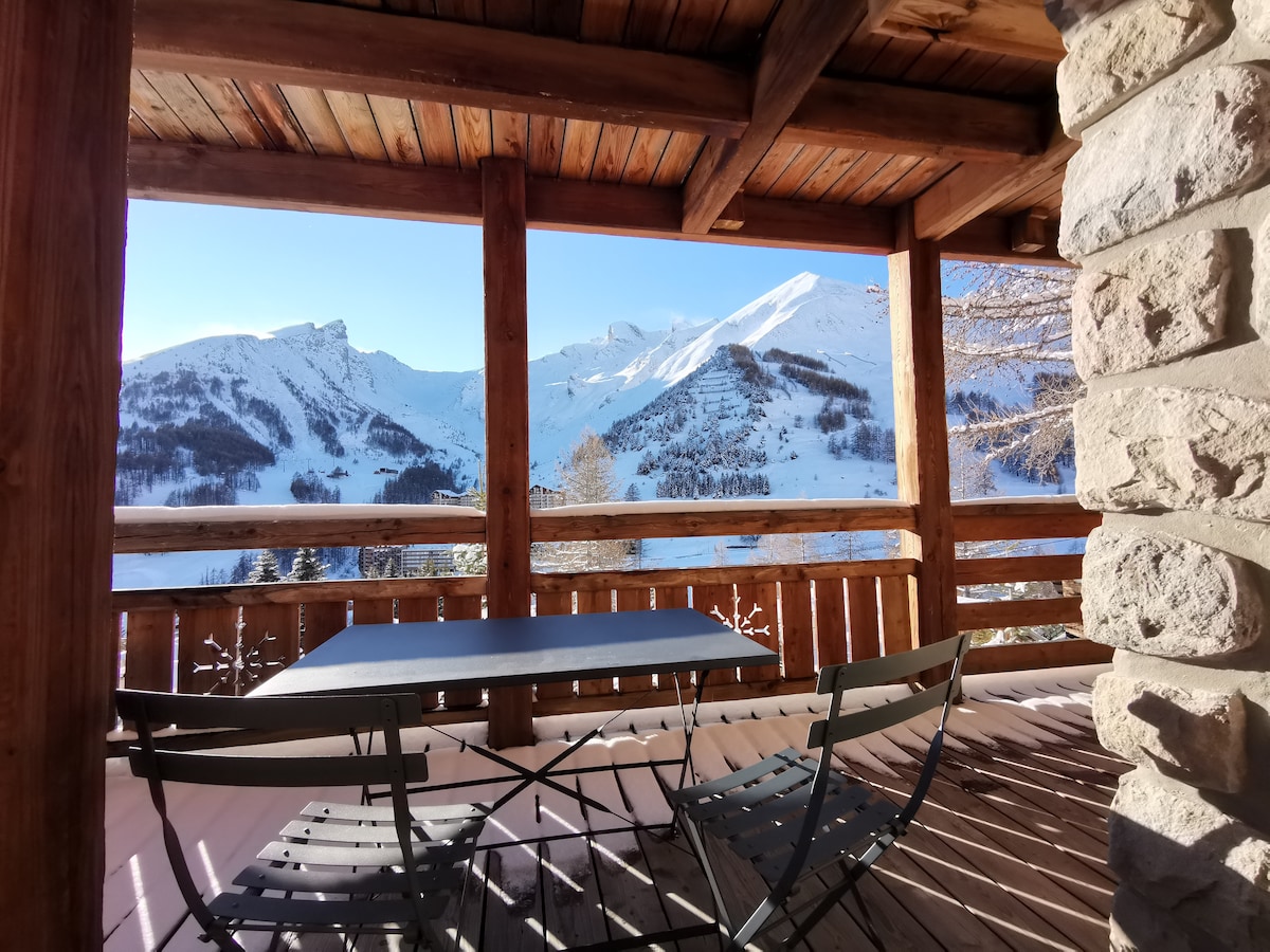 A terrace features a table and chairs, offering views of snow-covered mountains. The wooden structure includes logs and beams, enhancing the natural feel of the space. Sunlight illuminates the scene, creating a serene and inviting atmosphere.