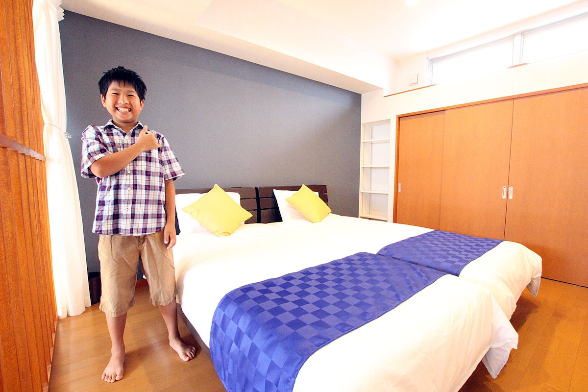 A well-lit bedroom features two spacious beds with blue and white bedding. Yellow decorative pillows add a touch of color. Natural light enters through large windows, illuminating the wooden floors and modern decor. A closet and shelves are visible in the background.
