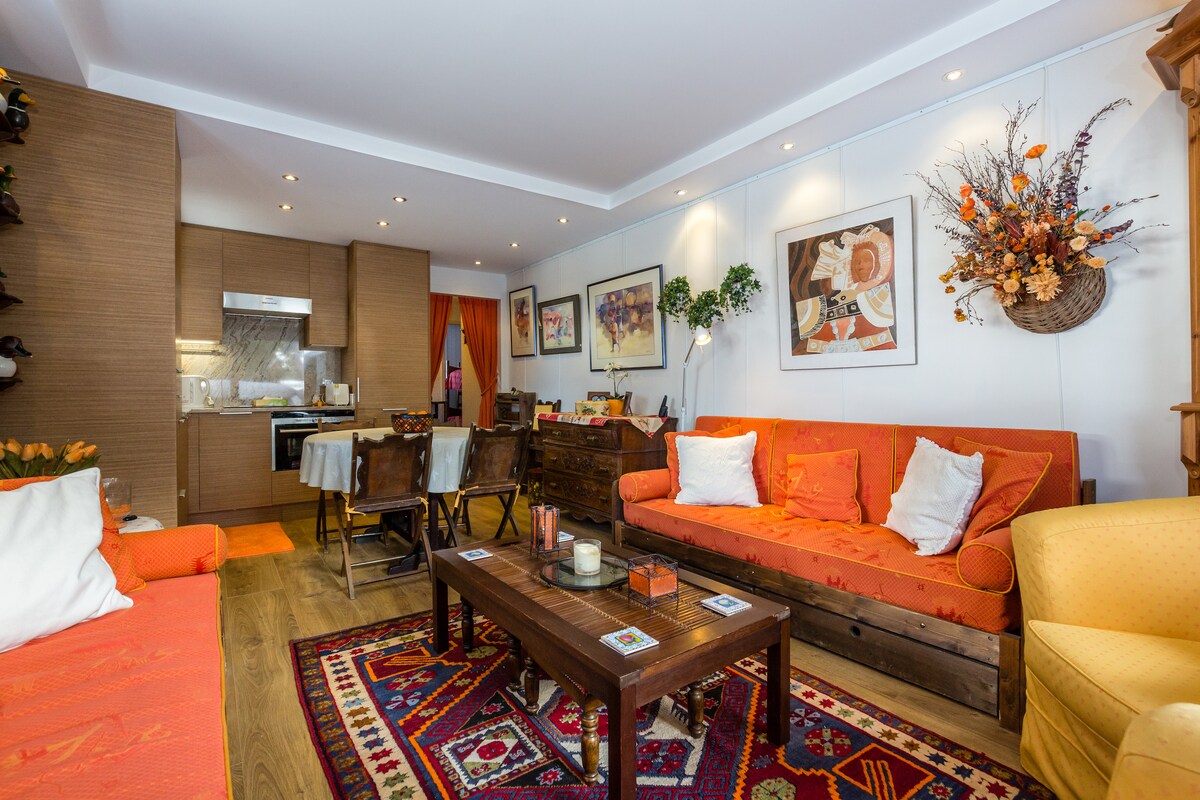 The open living space features warm, earthy tones with two inviting sofas arranged around a wooden coffee table. A spacious kitchen is visible, complemented by artful decor on the walls. Natural light brightens the area, enhancing the cozy ambiance.