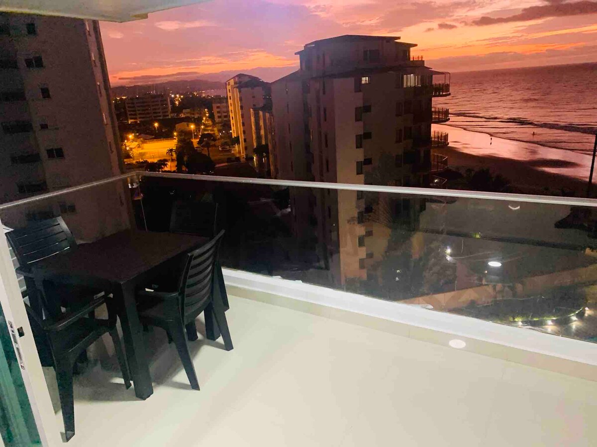 A spacious balcony features a dark dining table and chairs, with an expansive view of the sunset over the ocean. Nearby buildings are illuminated by city lights, and the shoreline is visible in the distance.