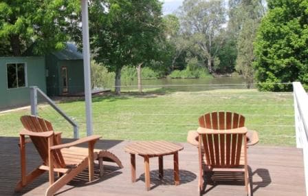 A spacious deck overlooks the Murray River, featuring two wooden adirondack chairs and a small circular table. Lush greenery and trees surround the water's edge, providing a tranquil setting for relaxation.