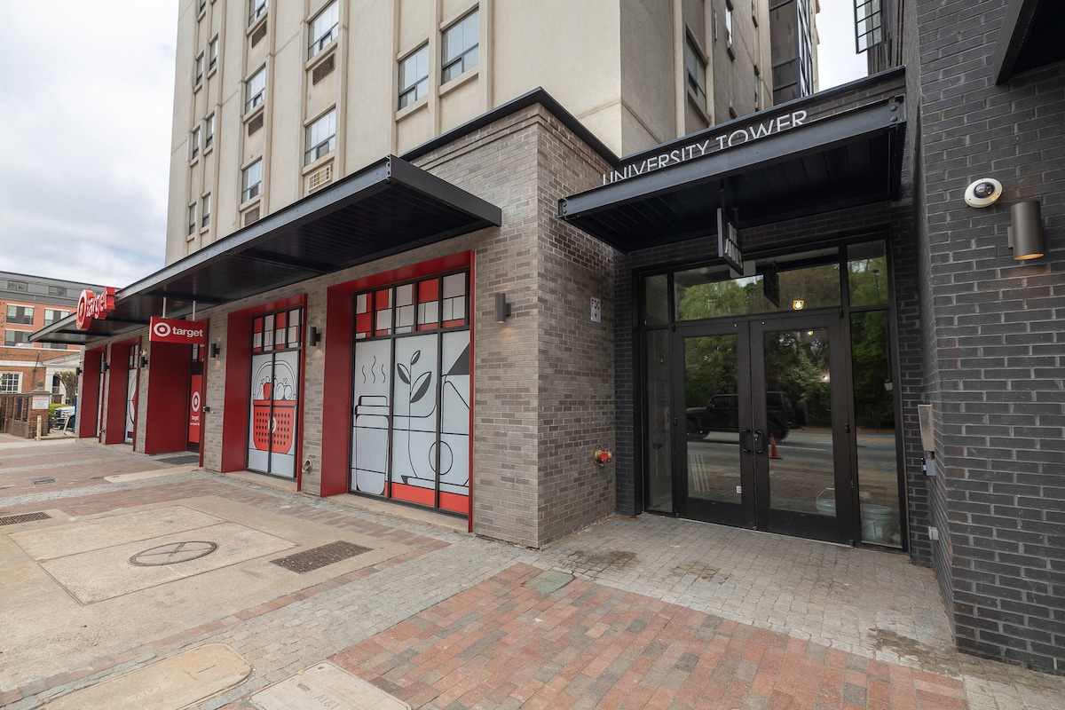 An urban building entrance is shown, featuring large glass doors with the name 'University Tower' prominently displayed above. The surrounding area includes storefronts with modern signage and a paved walkway, contributing to the downtown atmosphere.