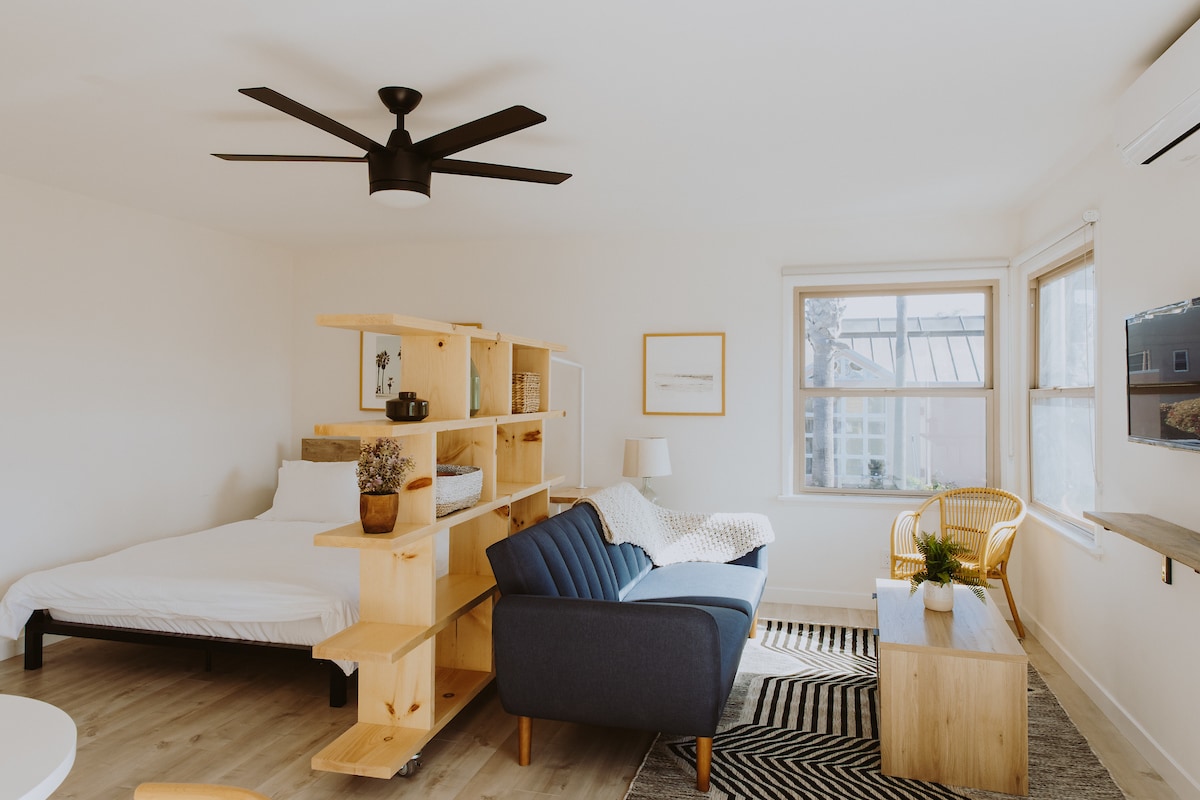 An open studio loft features a cozy layout with a queen bed, a stylish blue couch, and a simple wooden coffee table. A room divider with shelves adds a touch of modern design. Natural light enters through a large window, brightening the space.
