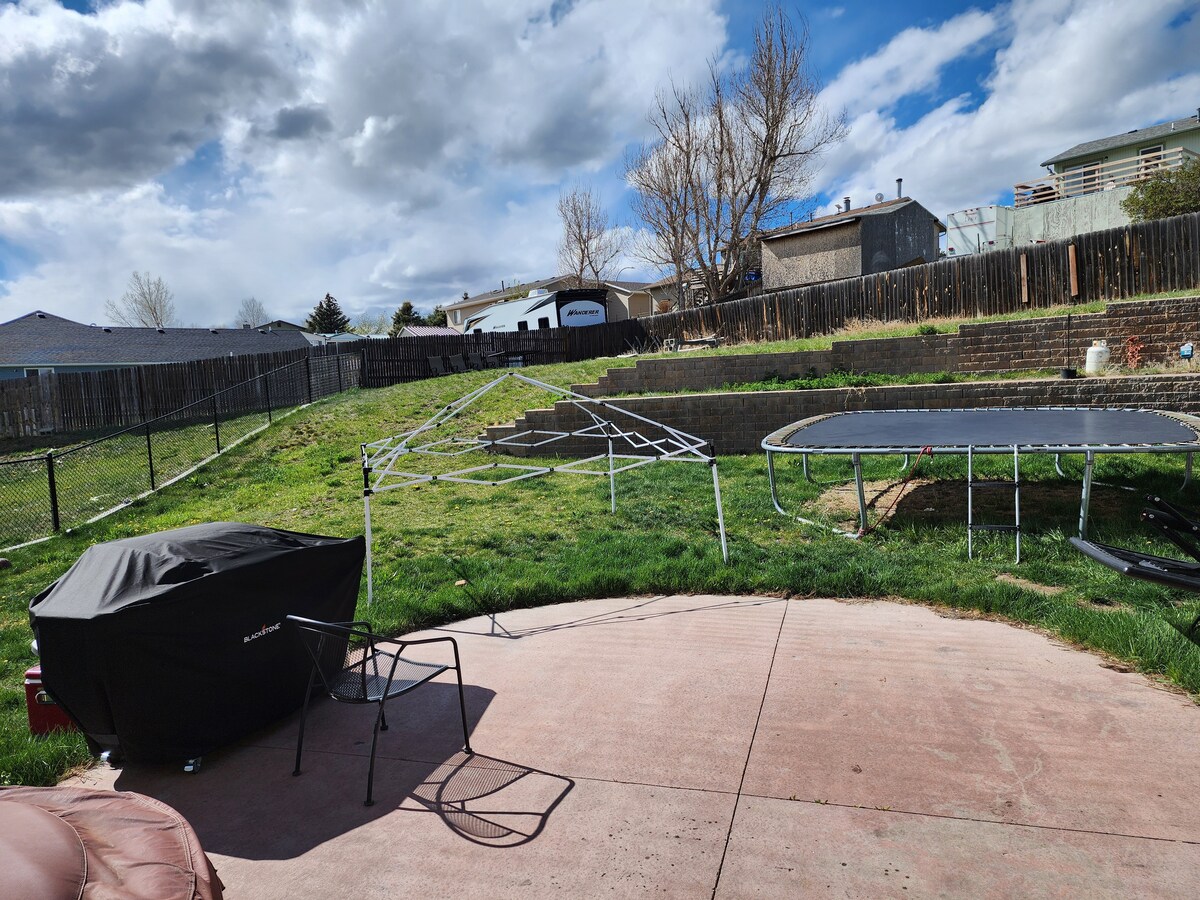 An open outdoor area features a grassy yard with a trampoline in view. A black cover is draped over a wooden structure, while a chair is positioned nearby. The landscape is framed by low fencing and a backdrop of residential buildings under a partly cloudy sky.