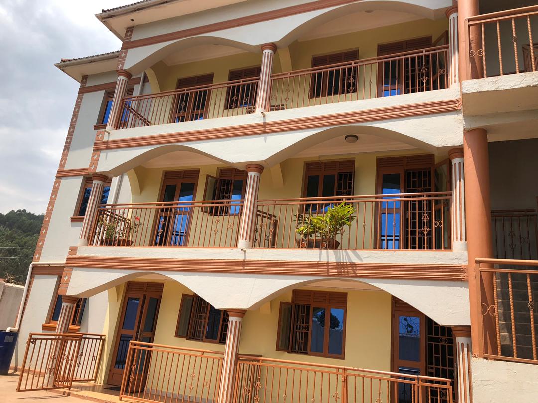 The exterior of the boutique hotel features a three-story structure with arched balconies and decorative columns. Potted plants are placed on some balconies, while large windows allow natural light into the interior. A serene environment is highlighted by the surrounding landscape.