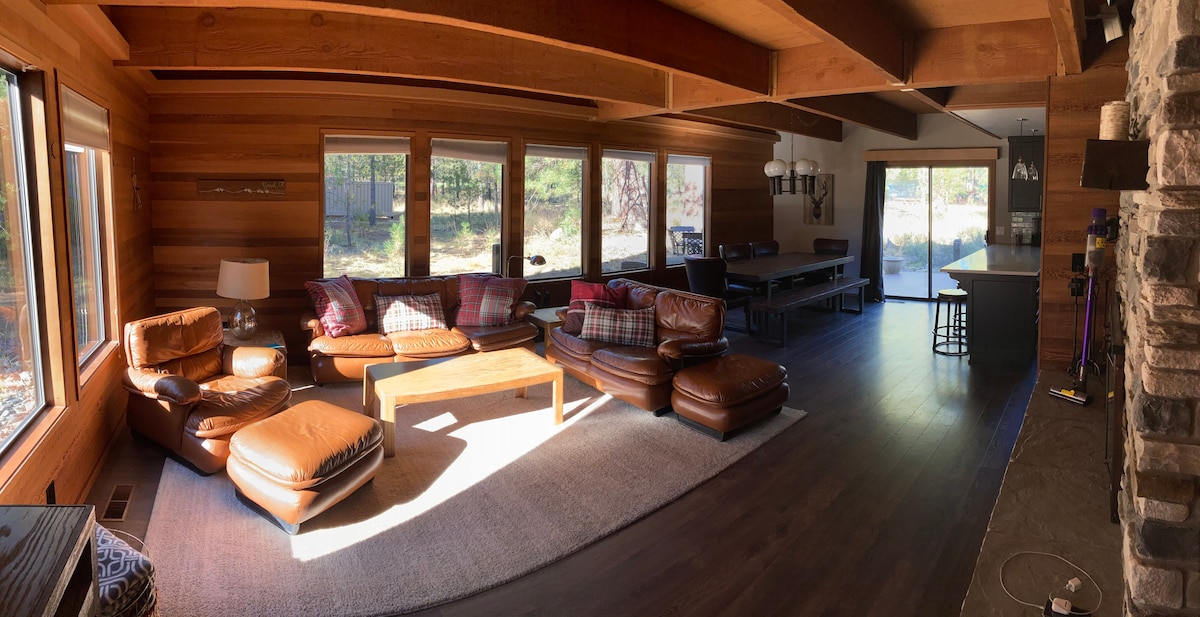 A spacious living area features large windows that allow natural light to fill the room. Comfortable leather seating is arranged around a wooden coffee table, with a cozy rug underneath. Visible in the background is a dining area and part of the kitchen, enhancing the open layout.