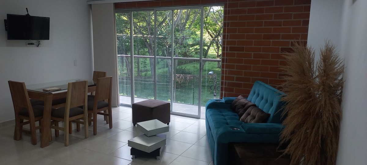 A modern living area features a turquoise sofa paired with a brown ottoman. A wooden dining table accommodates four chairs. Large glass doors open to views of surrounding greenery, while a wall-mounted television is visible above the dining area.