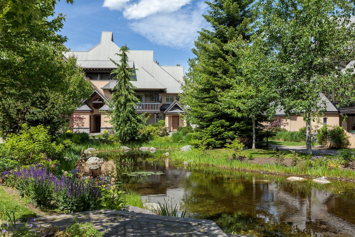 A serene outdoor area features a calm pond reflecting lush greenery and colorful flowers. Tree-dotted spaces create a natural ambiance, while charming architecture is visible in the background, enhancing the tranquility of the environment.