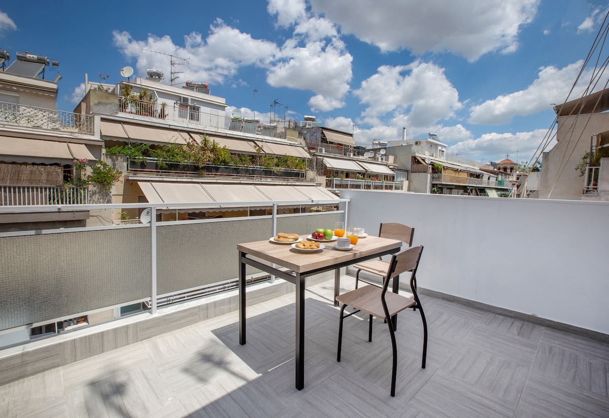 A sunlit veranda features a wooden table set for breakfast with various fruits and drinks. Surrounding buildings offer a glimpse of urban life, and the clear blue sky complements the inviting atmosphere.