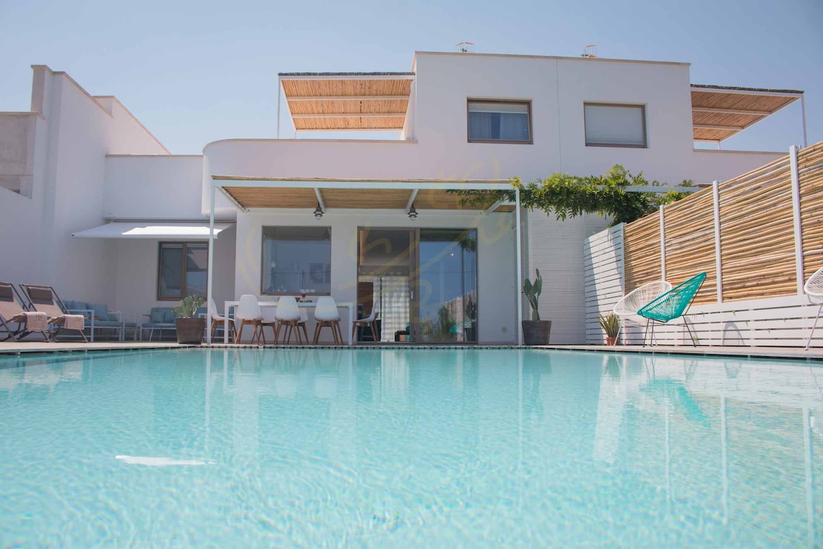 The image captures a modern villa with a clear blue pool reflecting the sky. A spacious terrace area features comfortable seating and a dining table. The house is surrounded by minimalistic landscaping, promoting a bright and airy atmosphere.