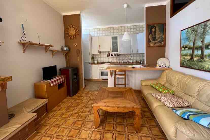 A living area features a light-colored sofa and a wooden coffee table positioned on a patterned floor. A small dining area with a bar counter and stools is visible. The kitchen is equipped with white cabinetry and appliances, while artwork decorates the walls.