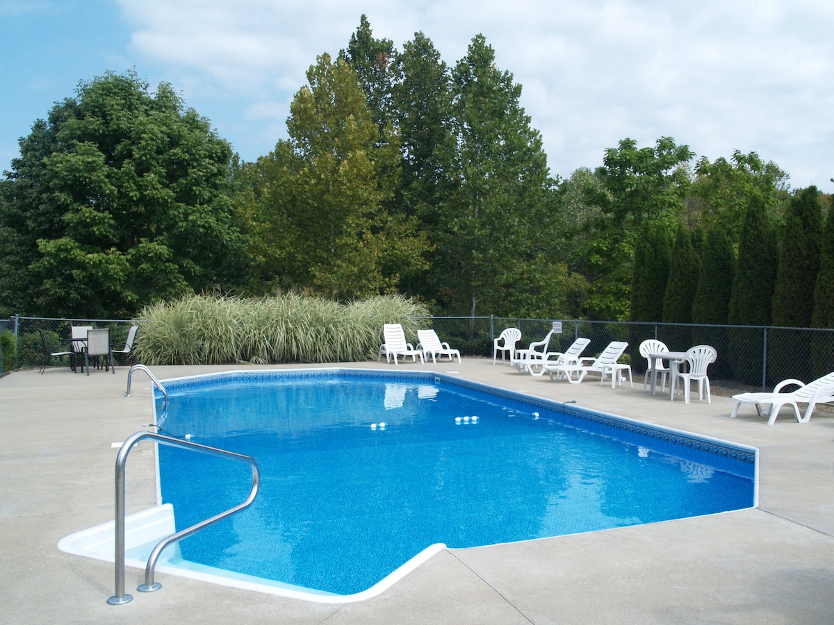A refreshing saltwater pool is surrounded by a spacious patio area, featuring lounge chairs and tables. Lush greenery and trees provide a natural backdrop, creating a relaxed outdoor setting. The water is clear and inviting, ideal for summer enjoyment.