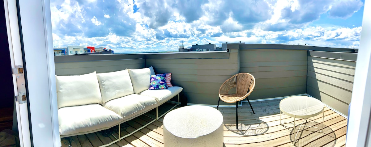 A rooftop balcony is furnished with a light-colored sectional couch and a round coffee table. A single armchair is positioned nearby. The space is surrounded by a contemporary railing, offering expansive views under a partly cloudy sky.