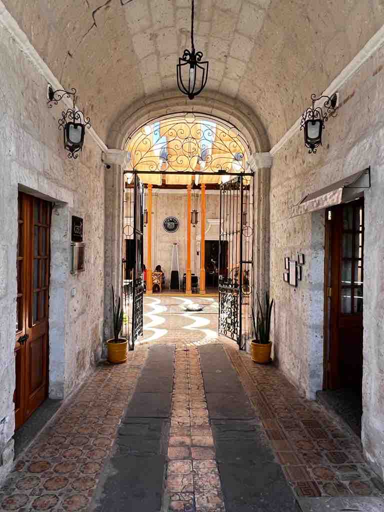 An arched entryway is framed by stone walls, leading to a vibrant courtyard. Intricate ironwork gates are adorned with lanterns, allowing natural light to illuminate the patterned tiles and lush plants positioned at the entrance.