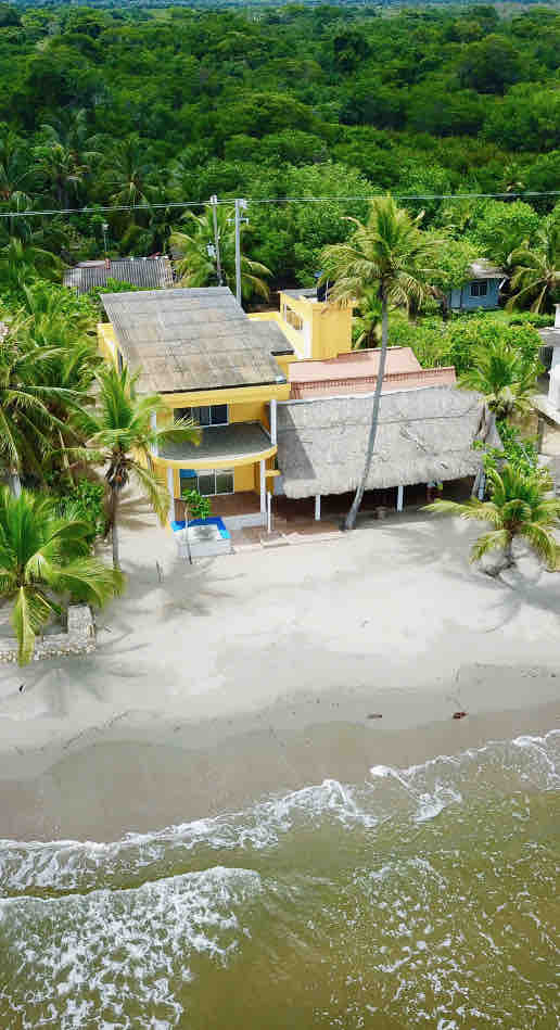 An aerial view of a beach house situated along the coastline, surrounded by lush greenery and palm trees. The structure features a sandy area leading to the water's edge and includes a thatched roof covering part of the entryway.