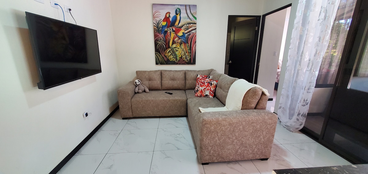 A spacious living area features a large, inviting sectional sofa adorned with colorful throw pillows. A mounted television is positioned on the wall, and a vibrant piece of wall art adds a touch of personality. Natural light flows in through large windows with sheer curtains.