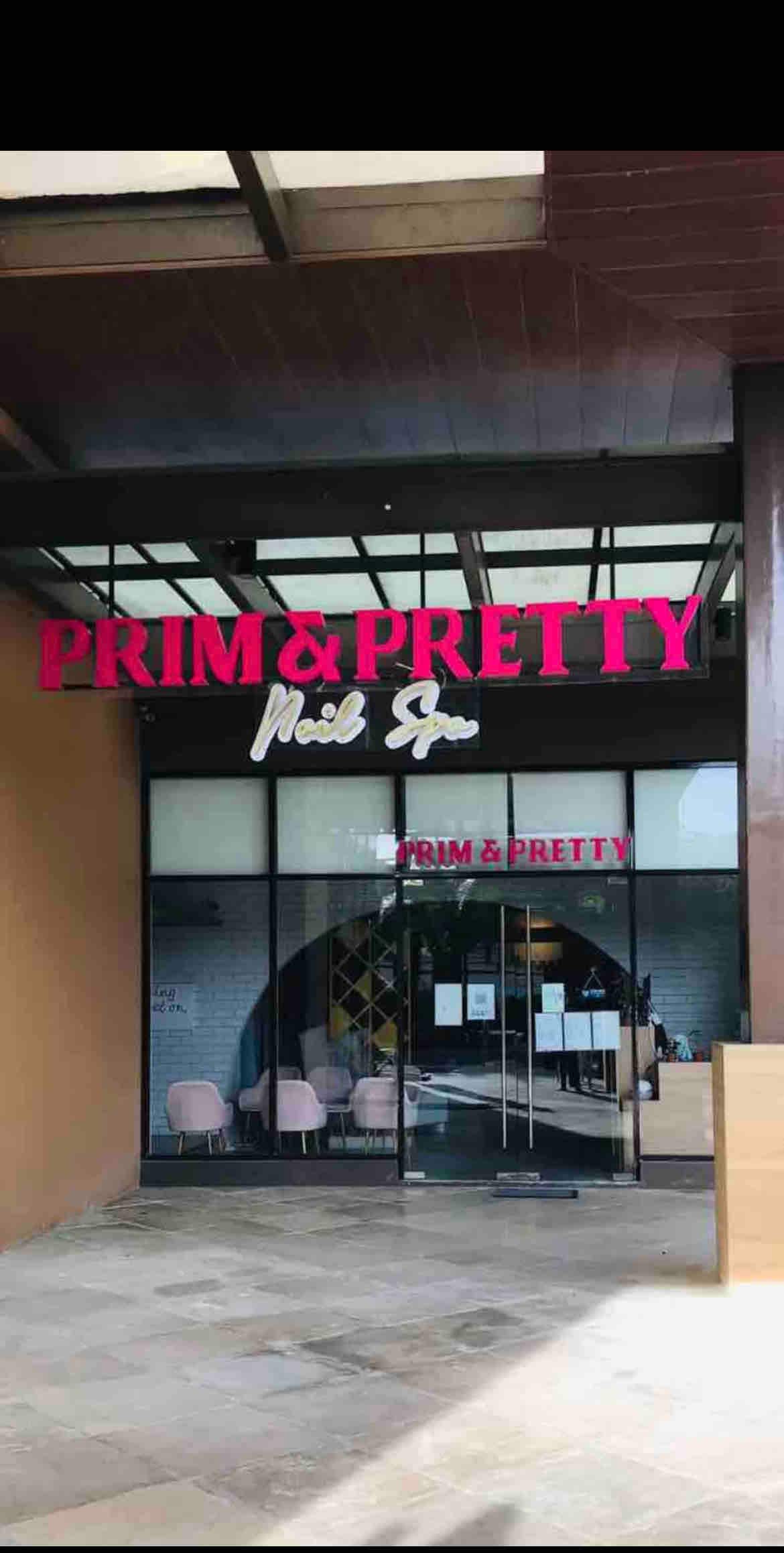 The exterior of a beauty salon features a glass entrance with large sliding doors. Above the entrance, a sign displays the name 'PRIM & PRETTY' in bold pink letters. The inviting space includes pale pink seating visible through the glass.