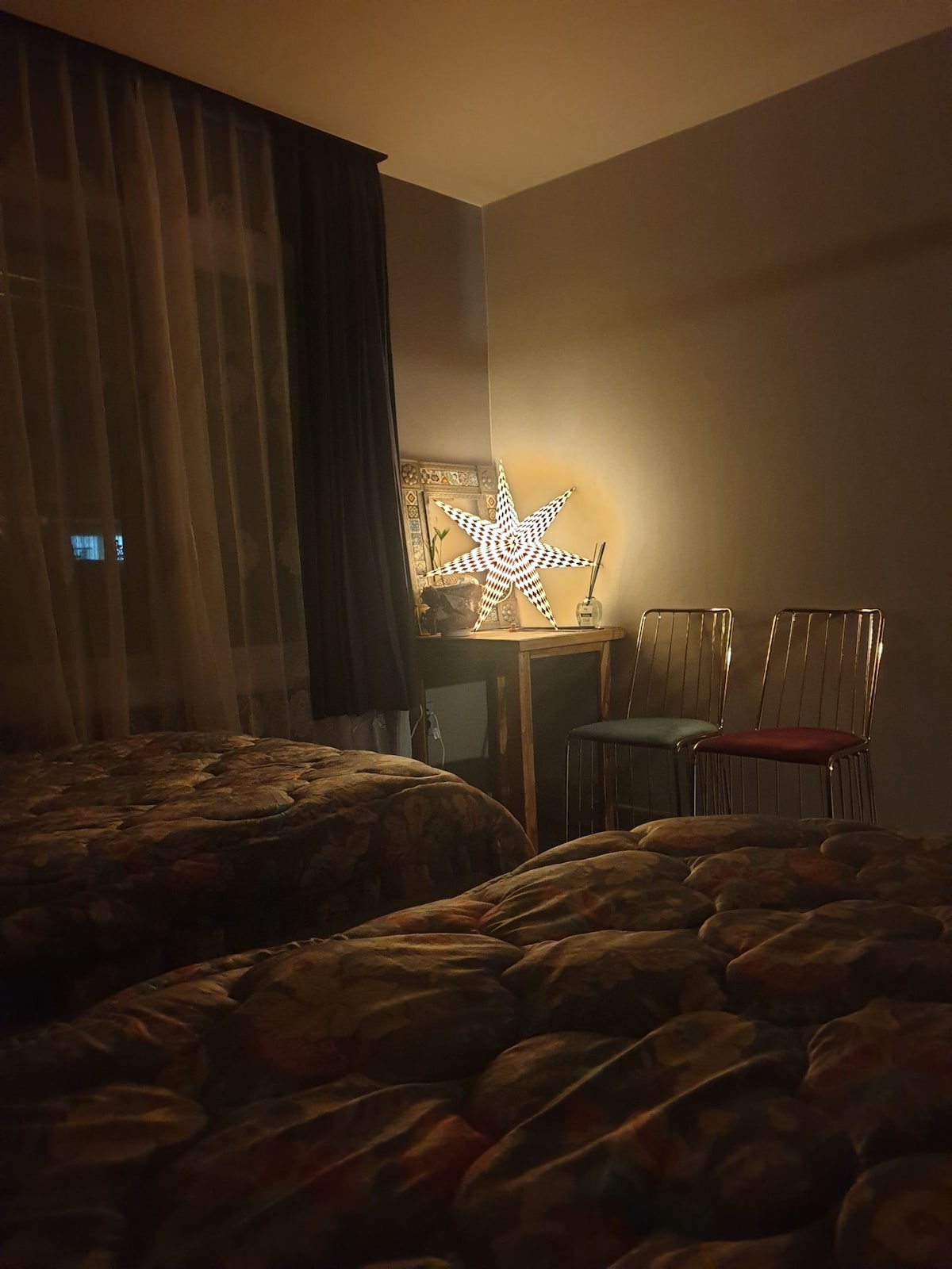 A softly lit guestroom features two spacious beds with patterned bedding. A small table holds a decorative star-shaped lamp, casting a warm glow. Two metal chairs are positioned nearby, and curtain drapes frame a window, creating a cozy atmosphere for relaxation.