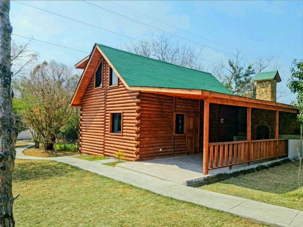 A charming log cabin features a green roof, surrounded by a grassy lawn and trees. A welcoming porch with wooden railings offers a space for relaxation. The driveway leads to the entrance, providing easy access to the cabin's outdoor area.