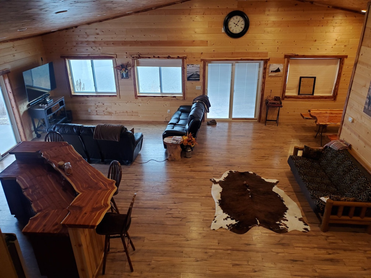 The spacious living area features natural wood paneling and large windows that allow for ample light. Two black sofas and a patterned futon are positioned around a decorative cowhide rug. A wooden bar with stools adds to the inviting space, complemented by a clock hung above.