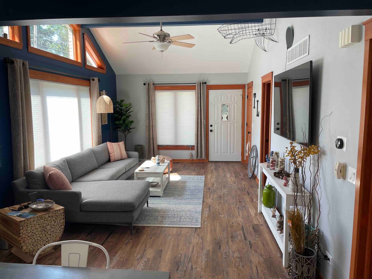 A bright living area features a spacious gray sectional sofa with colorful cushions. A light wooden coffee table sits in front of the sofa, complemented by a decorative rug. Large windows allow natural light to fill the room, enhancing the welcoming ambiance.