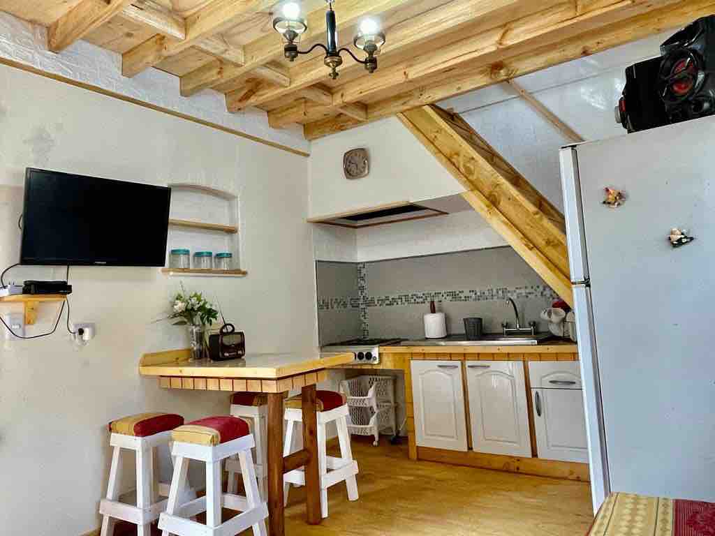A cozy kitchen area is visible, featuring a wood dining table with three stools and a flat-screen TV mounted on the wall. Kitchen appliances are neatly arranged, and wooden beams complement the space, enhancing the rustic charm.