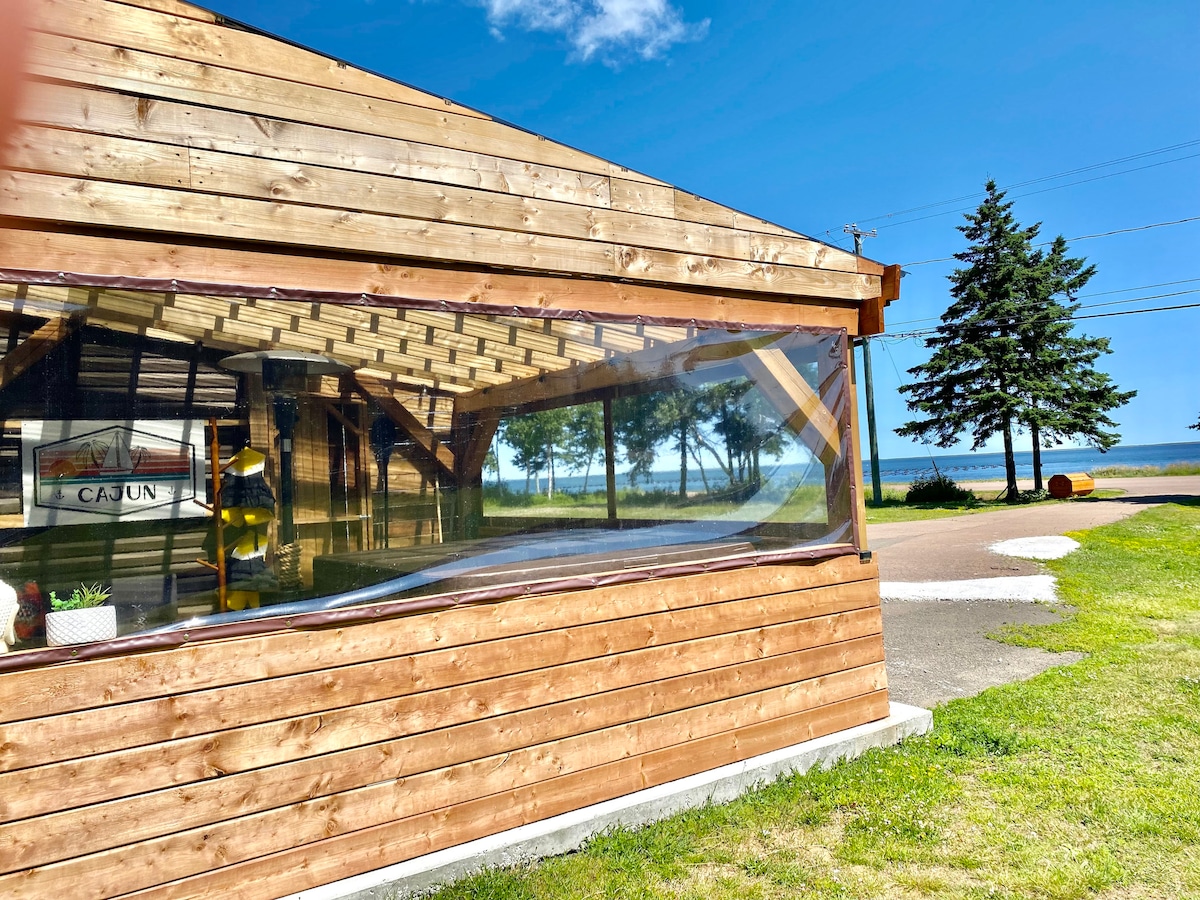 A wooden exterior features large glass windows that provide an unobstructed view of the ocean and surrounding landscape. The vibrant blue sky contrasts with the natural wood tones, enhancing the cottage's seaside charm. Lush greenery frames the view, creating a serene atmosphere.