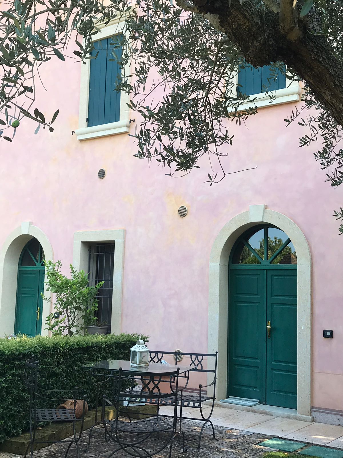 A charming exterior showcases a pale pink façade accented by two green doors and arched windows. A small outdoor seating area is visible, featuring a dark metal table surrounded by chairs, set beneath the shade of an olive tree.