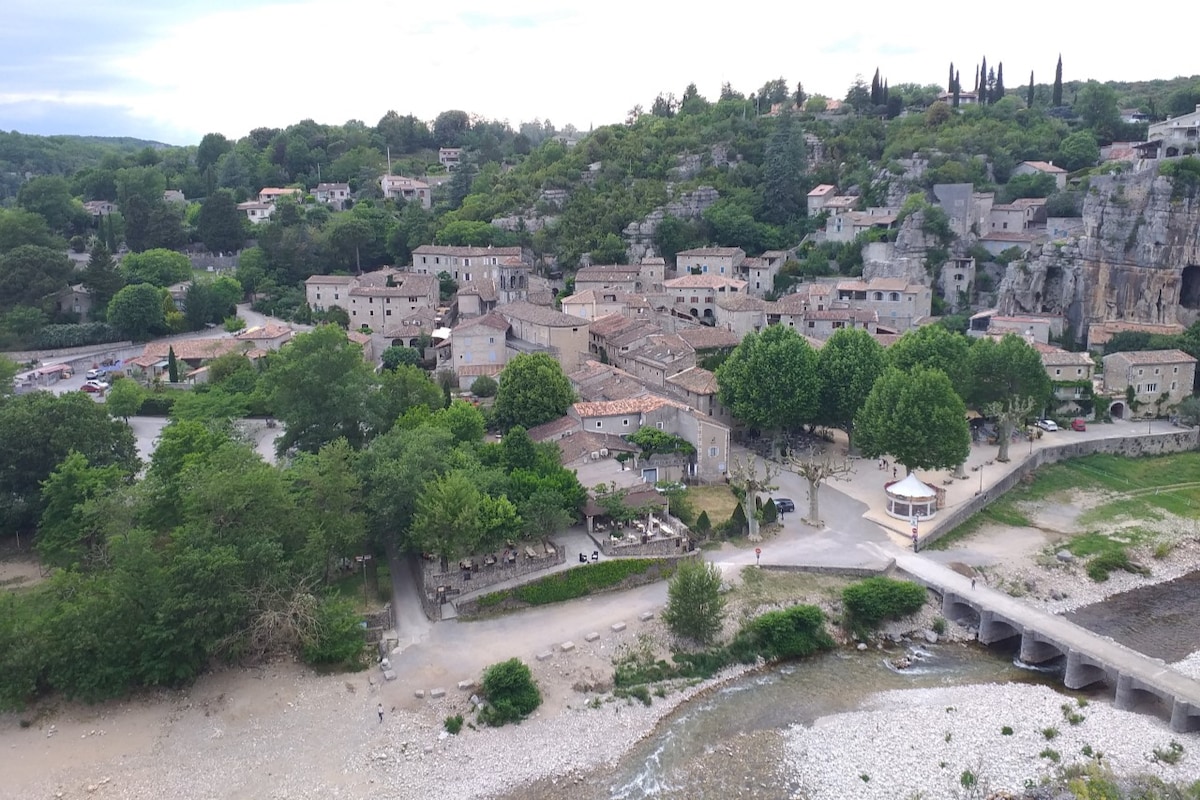 A picturesque view of a quaint village is presented, nestled among greenery and rocky terrain. Stone buildings with terracotta roofs are visible, along with trees lining the streets and a river flowing nearby. The scene captures a calm atmosphere in a natural setting.