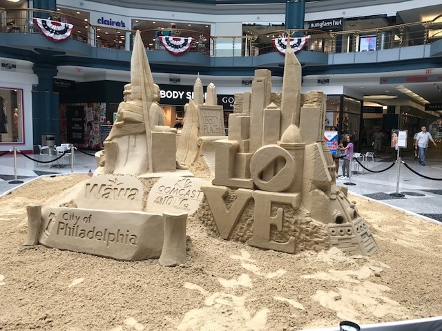 A large sand sculpture displays iconic Philadelphia landmarks, including the Liberty Bell and the Comcast Center, with the word 'LOVE' prominently featured. The artwork is surrounded by smooth sand, located within the spacious interior of a shopping mall, with shoppers visible in the background.