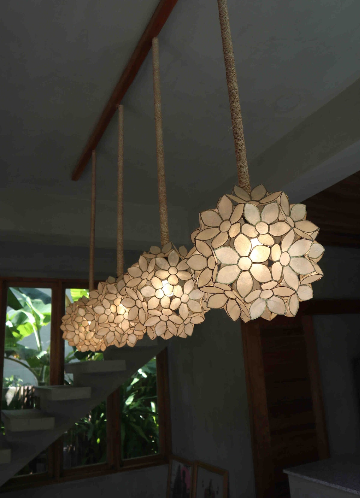 Three pendant lights hang from a wooden beam, featuring floral designs crafted from a light-colored material. The warm glow from each fixture enhances the surrounding space, illuminating the wall and staircase below. Green plants are subtly visible through large windows in the background.
