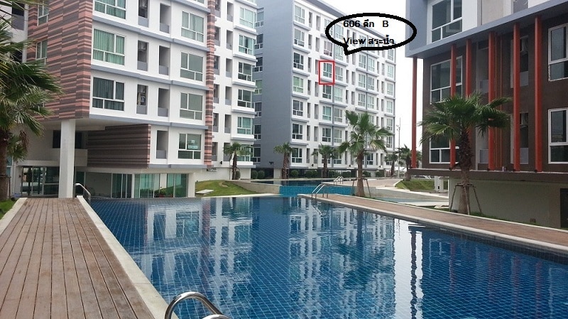 A well-maintained swimming pool is featured in the foreground, reflecting the surrounding buildings. The modern condominium structure showcases large windows and balconies, with green palm trees lining the walkways. The peaceful atmosphere is highlighted by well-manicured grass areas beside the pool.
