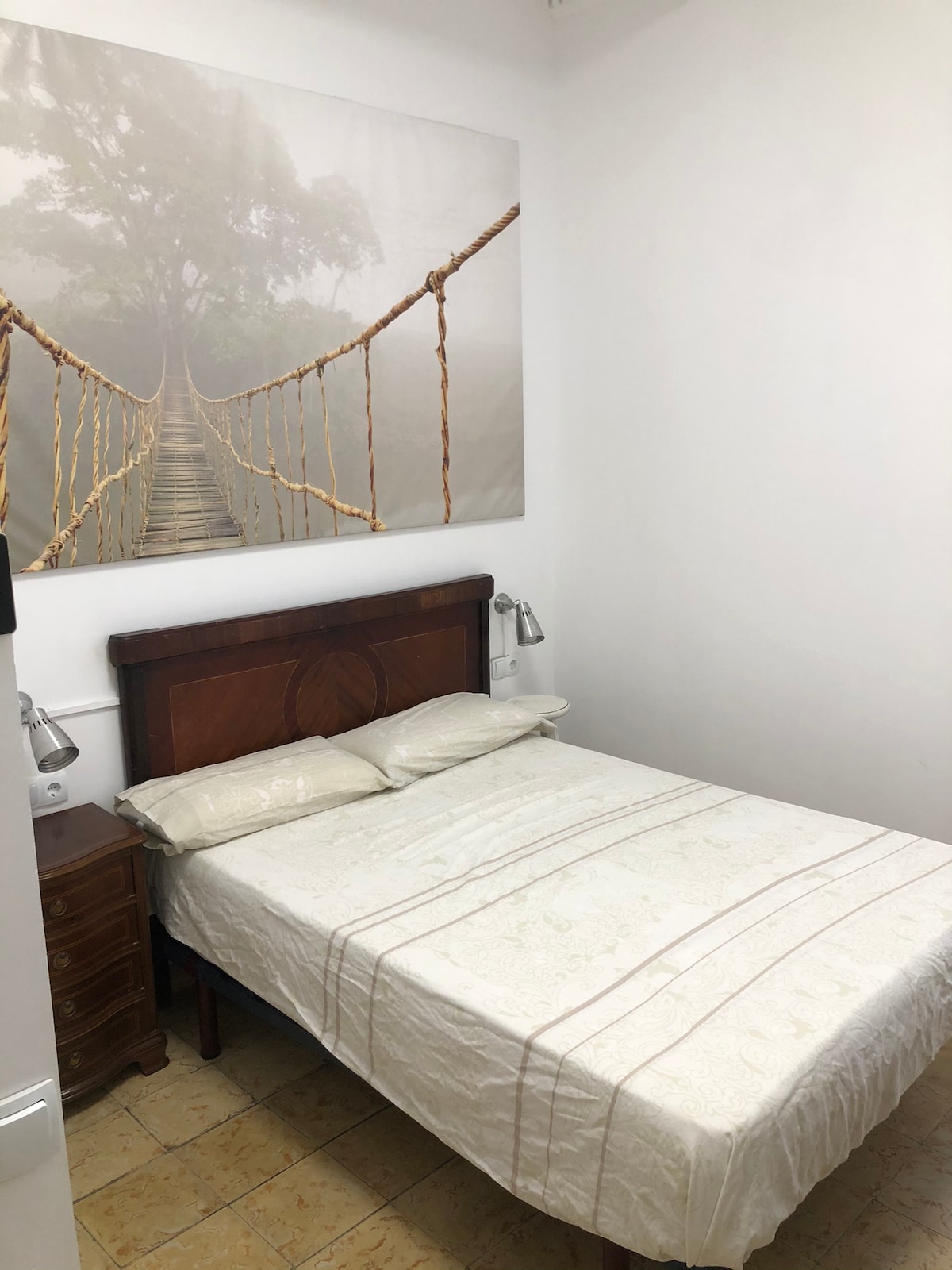 A private room features a spacious bed with light-colored linens, positioned against a wall adorned with a large, tranquil artwork of a bridge in mist. Two bedside lamps provide illumination, and a wooden nightstand is visible beside the bed.