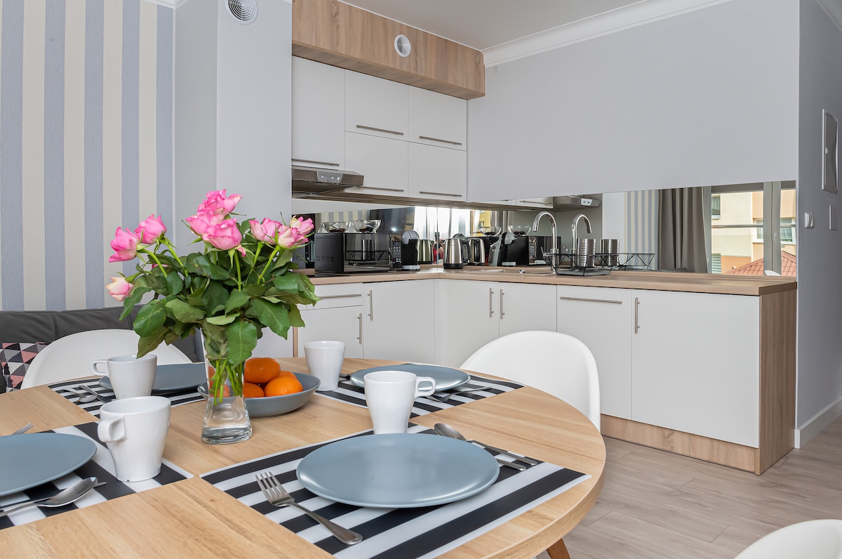 A dining area is set with plates and cups arranged around a light wooden table. A vase filled with pink flowers sits center-stage, while a kitchen area with modern appliances and sleek cabinetry is visible in the background.