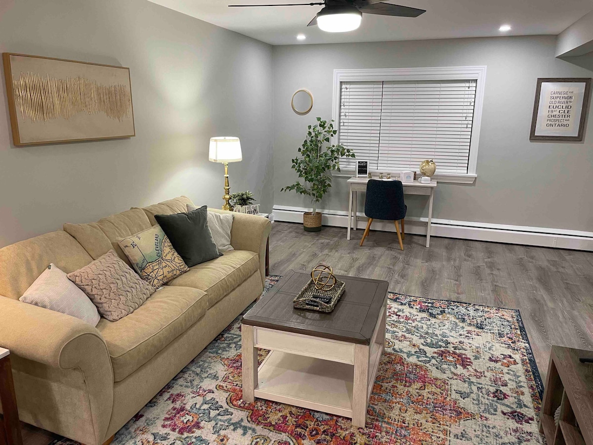 A cozy living room features a light-colored sofa adorned with decorative pillows. A coffee table sits center stage on a colorful area rug. A desk for work is positioned near the window, complemented by a plant and stylish lighting.