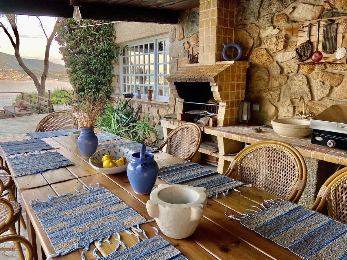 An outdoor dining area is presented with a large wooden table surrounded by woven chairs. A barbecue station is visible, made of stone, complemented by a tiled backsplash. Vases with fruits sit on the table, framed by greenery and a view of the water beyond.