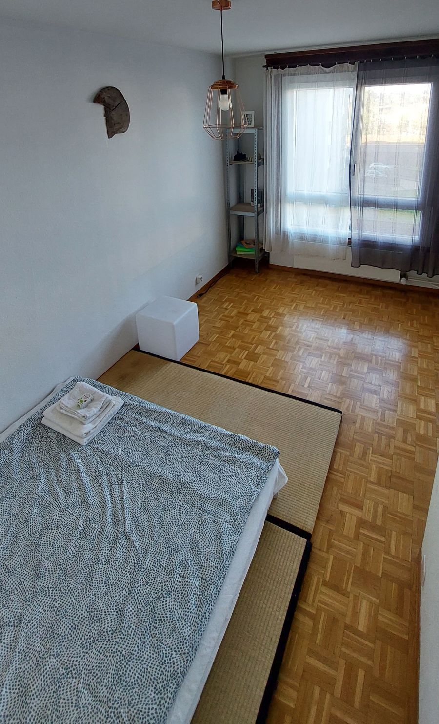 A spacious room features a low bed with a patterned blanket laid neatly. A bedside table with stacked towels sits nearby. Natural light streams in through large windows, which are partially covered by sheer curtains, illuminating the wooden flooring.