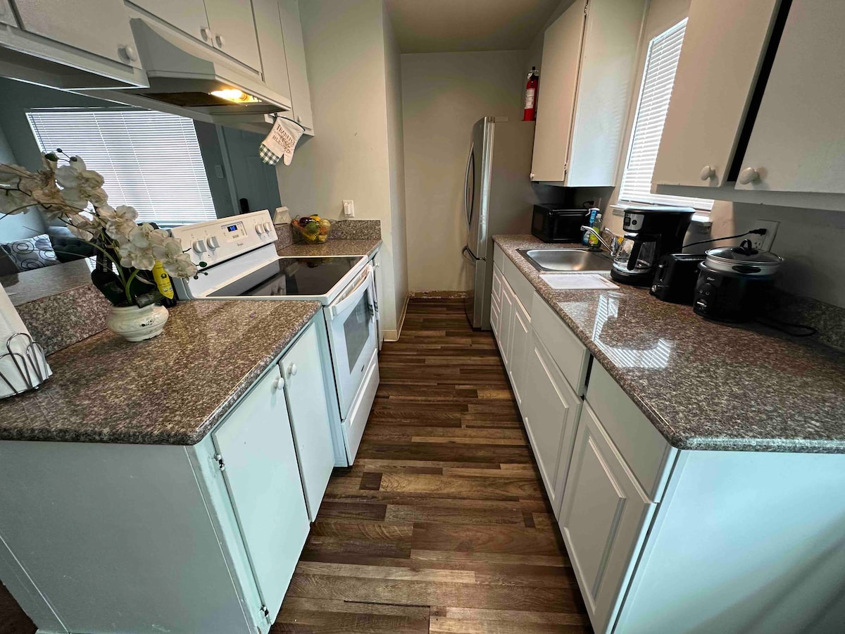 The kitchen features white cabinetry and granite countertops, complemented by modern appliances. A stainless steel fridge stands alongside an oven and microwave. Natural light enters through a nearby window, illuminating the space, which includes a small coffee maker and other essentials.