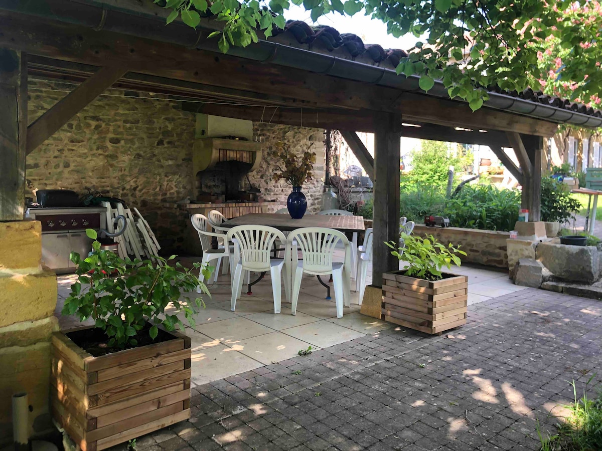 An outdoor covered barbecue area features a large table surrounded by white chairs, with stone walls providing a rustic backdrop. Potted plants add greenery, while the open space invites relaxation and dining in nature.