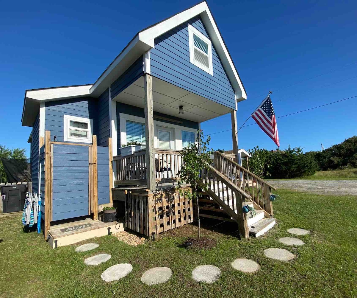A charming tiny home is displayed with a blue exterior and a white gabled roof. An American flag gently flutters in the breeze. Steps lead to a welcoming porch, surrounded by a landscaped yard featuring stepping stones and greenery.