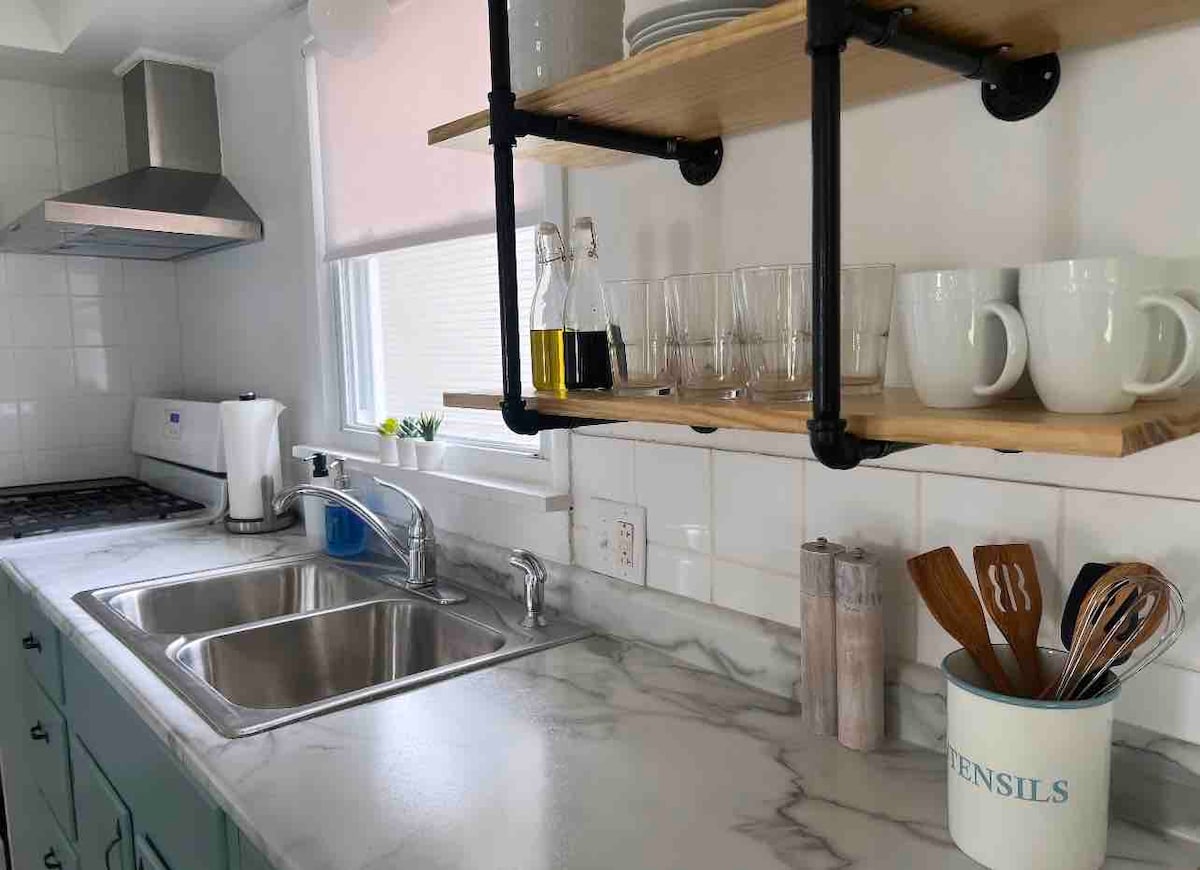 The kitchen features a modern design with a marble countertop and a stainless steel sink. Above, a wooden shelf displays glassware and cooking utensils. Appliances include a stove and a kettle, while natural light fills the space through nearby windows.