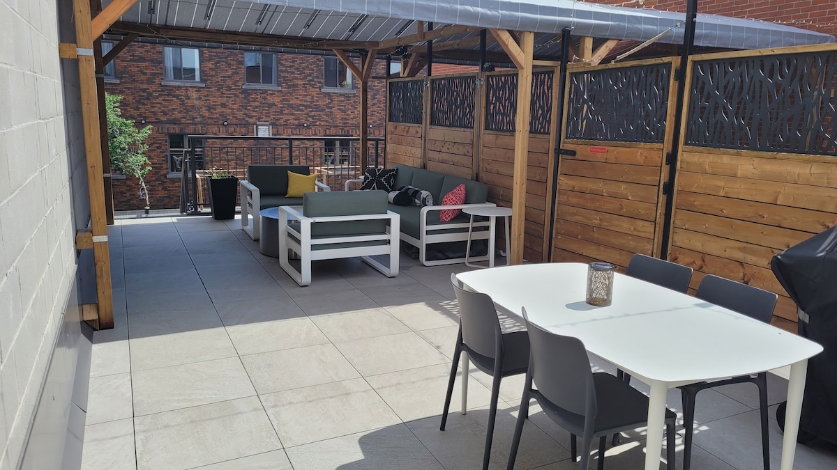A private terrace features a spacious dining table with six chairs and a lounging area with a sectional sofa and cushions. A Gaz BBQ is visible in the corner, offering an ideal space for outdoor relaxation and dining.