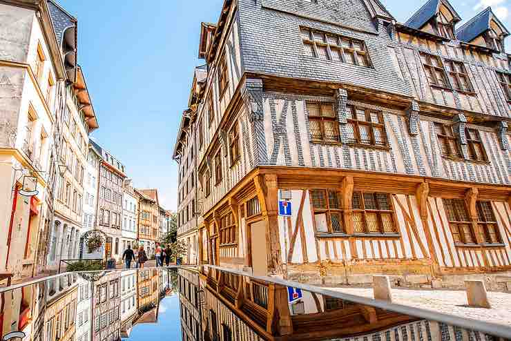 The image captures charming half-timbered buildings lining a cobblestone street, creating a historic ambiance. The scene reflects warm colors and intricate architectural details, while the sky is clear and bright. People can be seen strolling leisurely, adding life to the picturesque setting.