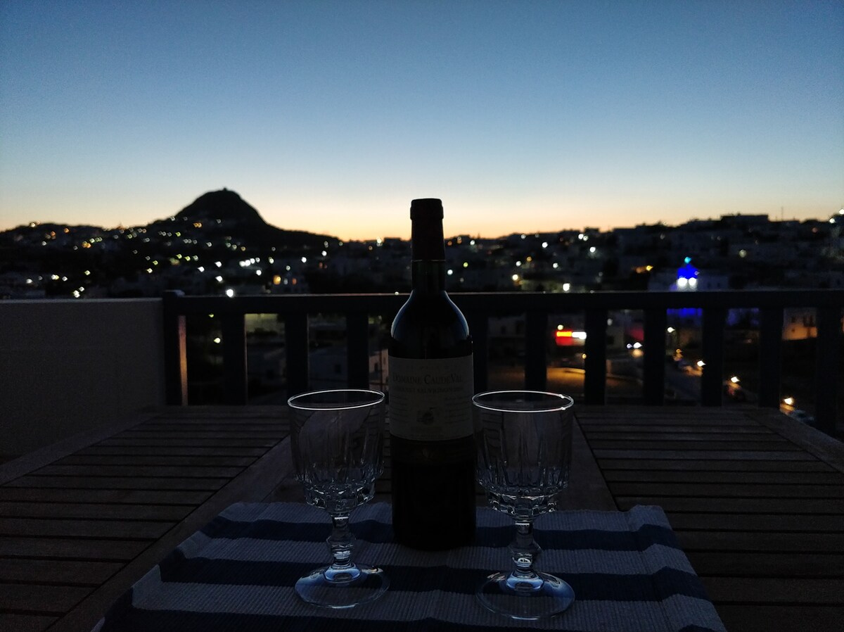 A wooden table is set for two with a bottle of wine and two crystal glasses. The backdrop features a twilight sky and distant hills illuminated by city lights, creating a serene atmosphere for relaxation.